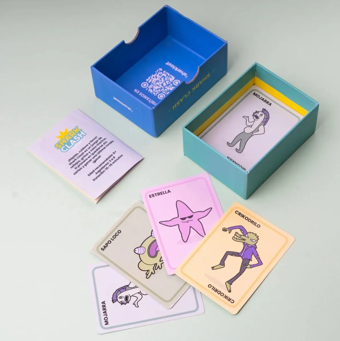 Caja abierta del juego Shark Clash con instructivo y cartas ilustradas de colores desplegadas alrededor.