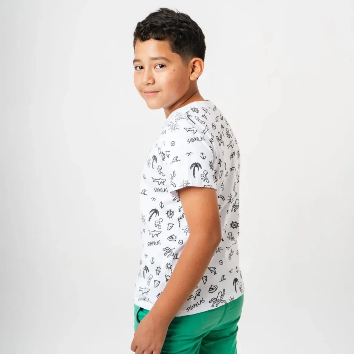 Niño de perfil con camiseta blanca de patrón ilustrado repetido y shorts verdes.