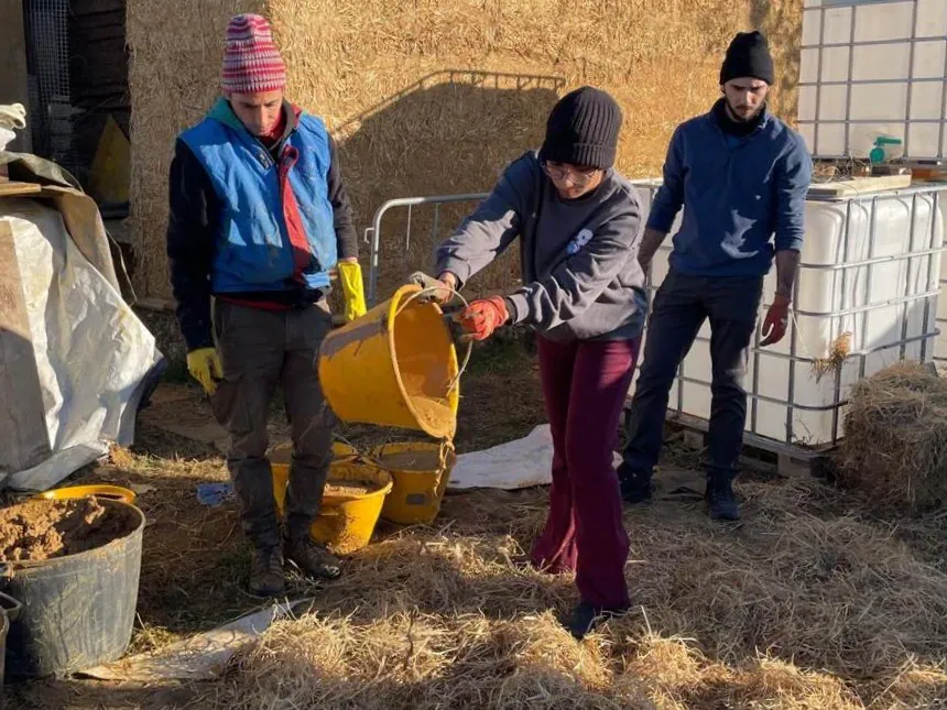 Tres personas abrigadas mezclando barro con paja en cubetas, al aire libre durante el invierno.